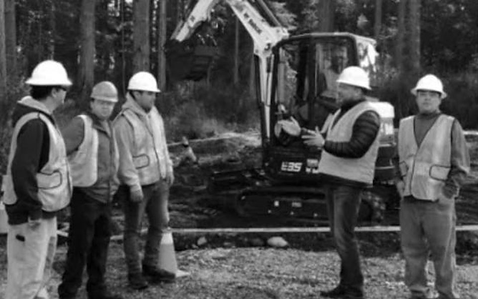 Grey Snow Training Academy instructor teaching trainees in hard hats beside a compact excavator